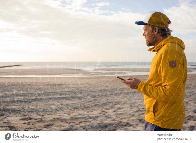 Man in yellow jacket, using smartphone on the beach use coat coats jackets Smartphone iPhone Smartphones beaches man men males mobile phone mobiles