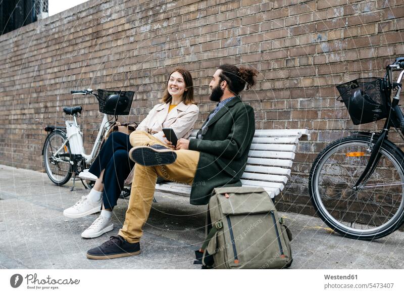 Couple sitting on a bench next to e-bikes talking Seated E-Bike Electric bicycle Electric Bike couple twosomes partnership couples bicycles speaking people