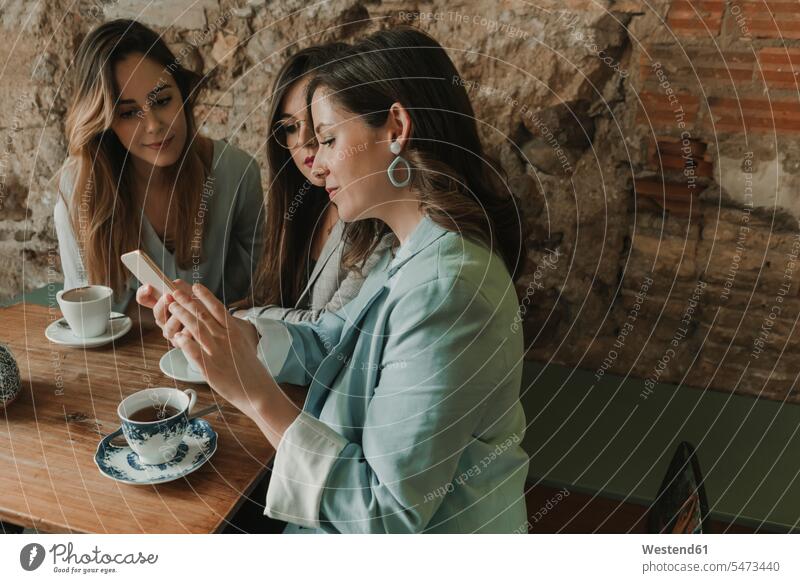 Three young women with a cell phone in a cafe Spain brick wall text messaging SMS Text Message brick walls fashionable message rustic brickwork stonework