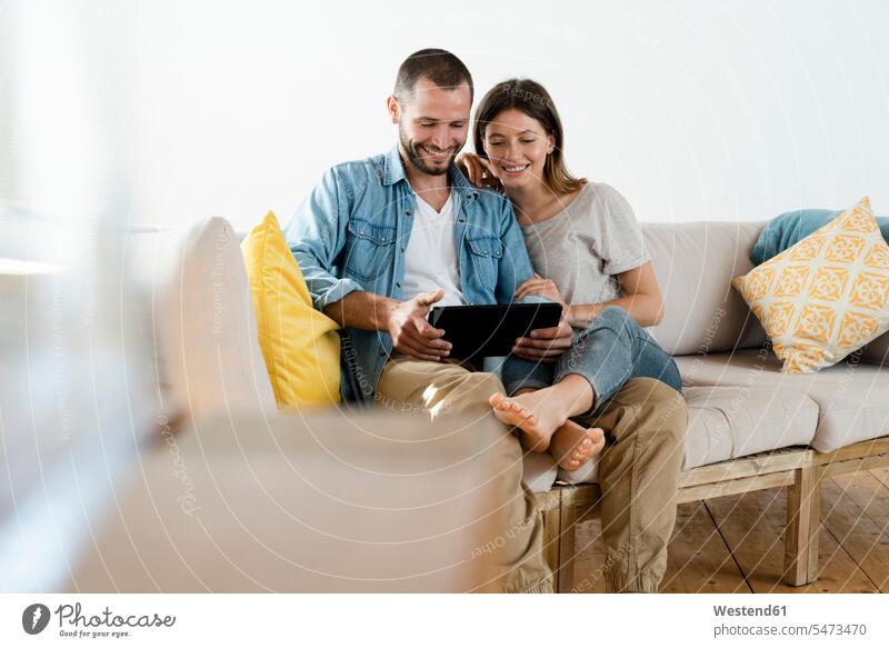 Happy couple at home in modern living room sitting on couch while looking at tablet together cushions couches settee settees sofa sofas hold smile Seated relax
