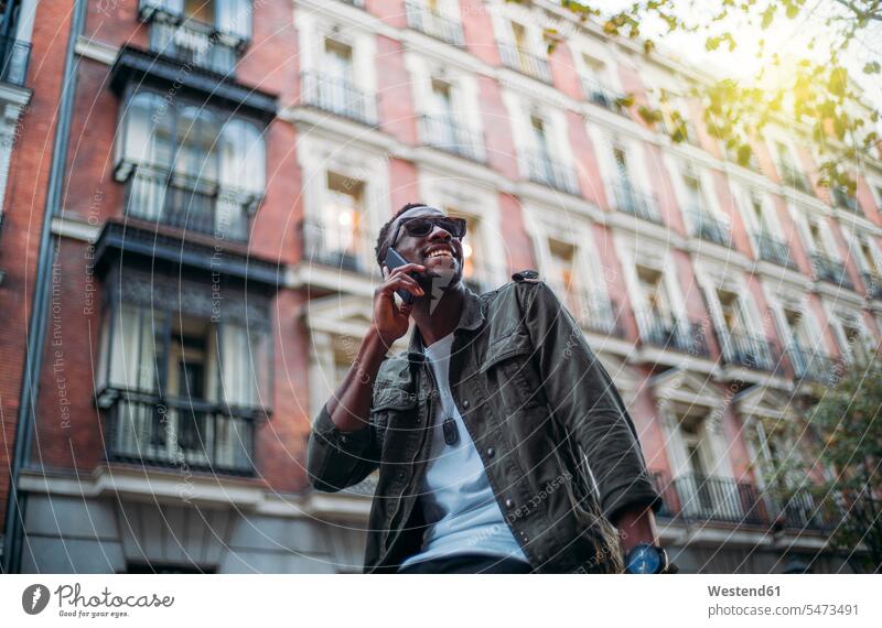 Smiling man wearing sunglasses talking over smart phone while sitting against building in city color image colour image Spain leisure activity