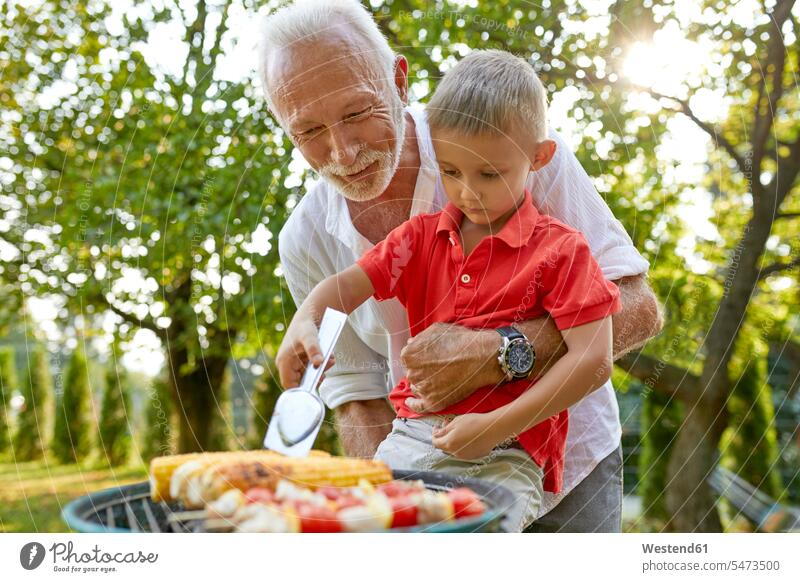 Grandfather helping grandson turning a corn cob during a barbecue in garden gardens domestic garden Barbecue BBQ Barbeque assistance assisting Help corncob