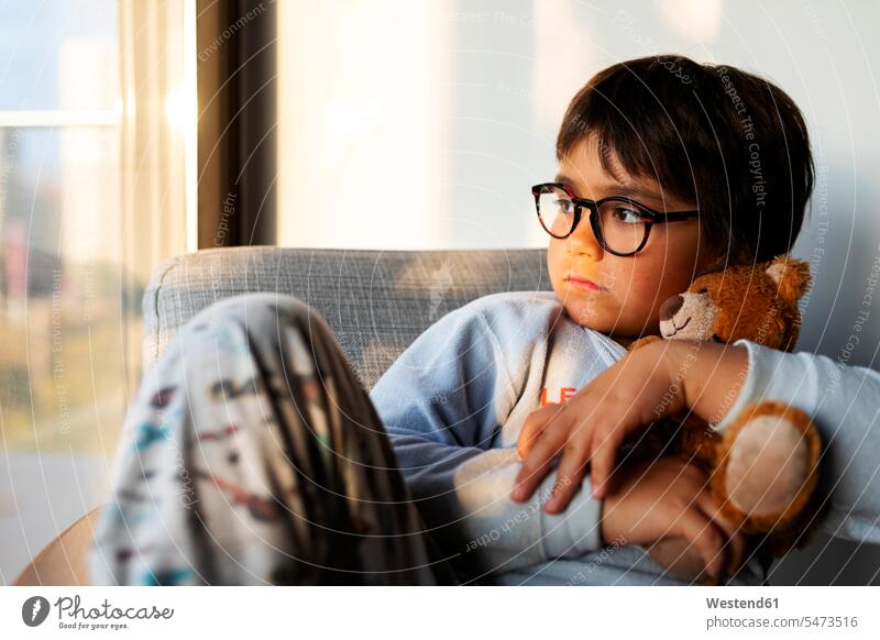 Portrait of serious boy with teddy bear sitting on armchair at home looking out of window windows pane panes window glass window glasses Window Pane windowpanes