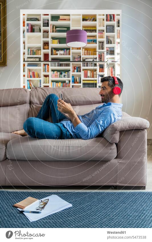 Smiling young man lying on the couch at home using smartphone and wireless headphones human human being human beings humans person persons caucasian appearance