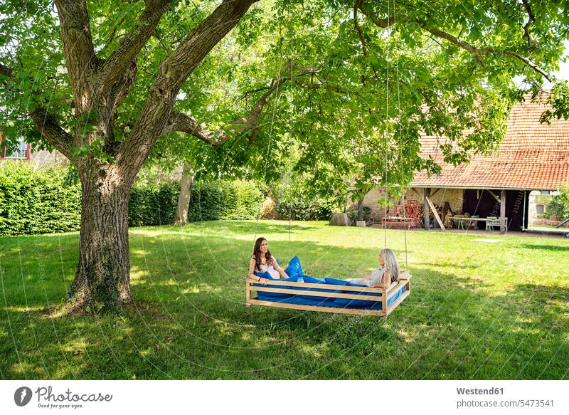 Two women relaxing on a hanging bed in garden beds relaxed relaxation female friends hammock hammocks woman females gardens domestic garden mate friendship