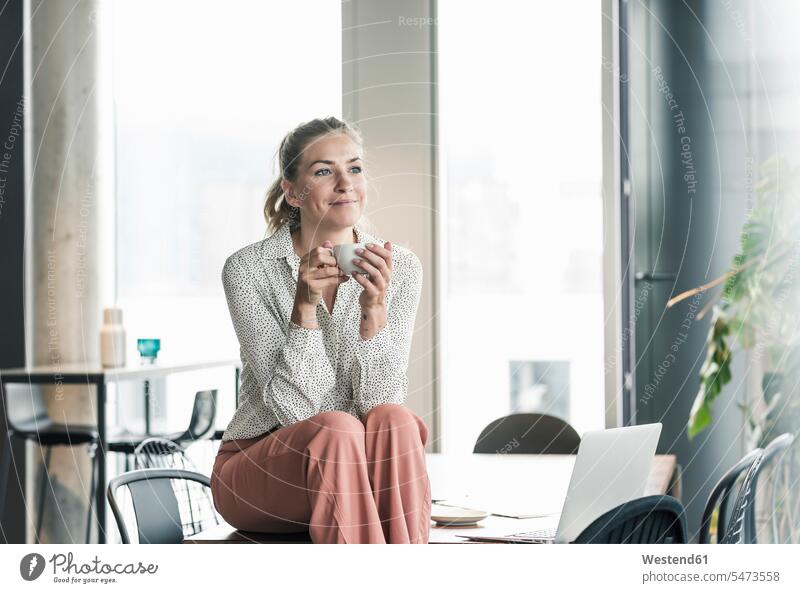 Smiling businesswoman sitting on table in office having a coffee break human human being human beings humans person persons caucasian appearance