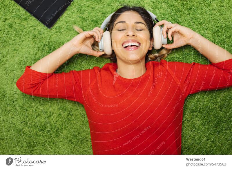 Happy young woman lying on carpet listening to music with headphones hearing happiness happy females women carpets rug rugs laying down lie lying down headset