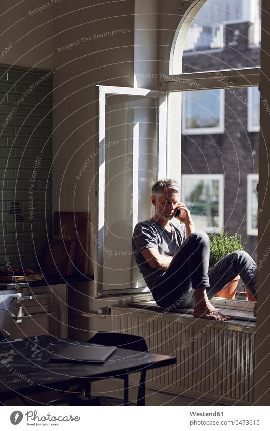 Mature man sitting at the window at home talking on the phone human human being human beings humans person persons celibate celibates singles solitary people