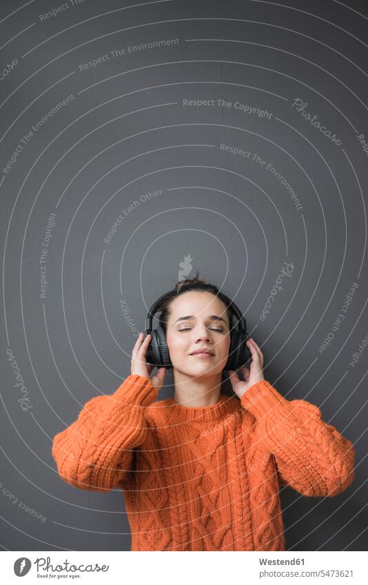 Portrait of woman wearing orange knit pullover leaning against grey wall listening music with headphones females women portrait portraits headset hearing walls