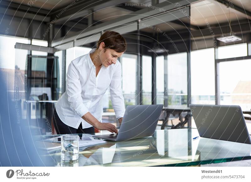 Businesswoman standing at glass table in office using laptop businesswoman businesswomen business woman business women offices office room office rooms