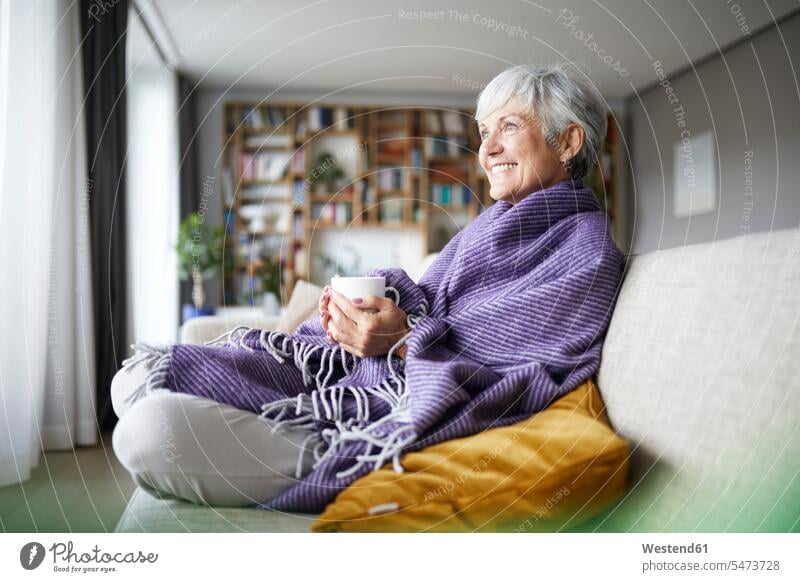 Smiling senior woman with blanket holding coffee cup while sitting on sofa at home color image colour image indoors indoor shot indoor shots interior