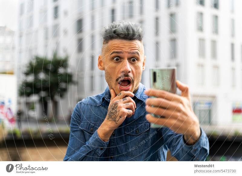 Shocked man holding smart phone during video call against building color image colour image Spain outdoors location shots outdoor shot outdoor shots day
