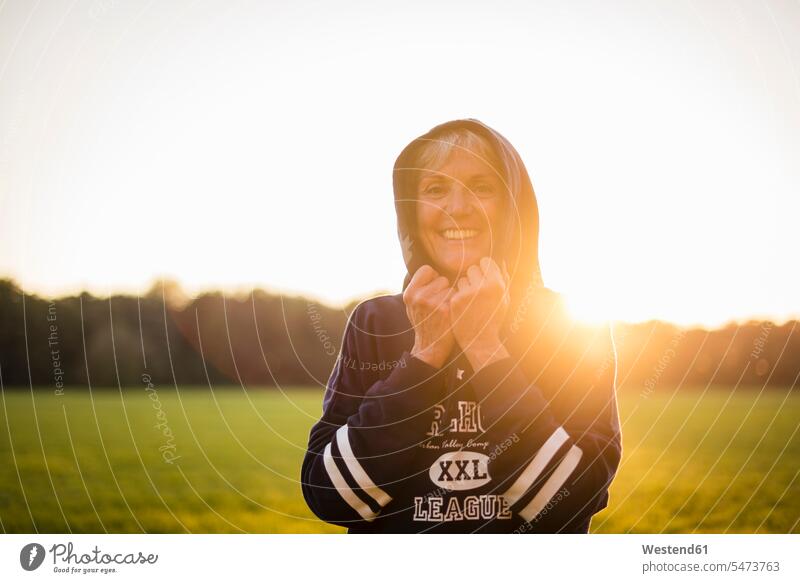 Portrait of smiling senior woman wearing a hoodie standing on rural meadow at sunset portrait portraits Hoodie Hoody sunsets sundown females women senior women