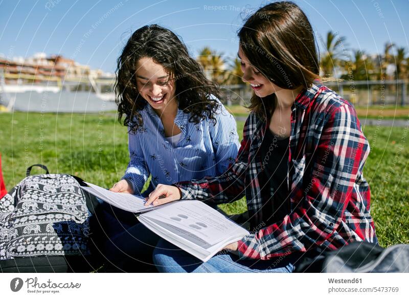 Two smiling female students sitting in park learning together parks smile Seated higher education discussing discussion backpack rucksacks backpacks back-packs