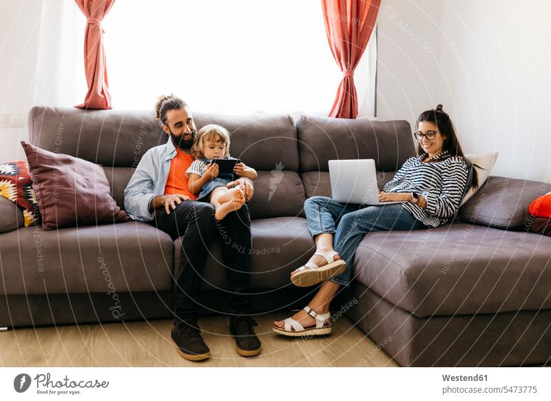 Woman working over laptop while father and daughter using mobile phone on sofa in living room color image colour image indoors indoor shot indoor shots interior