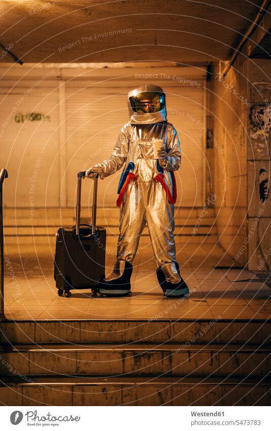 Spaceman in the city at night standing in underpass with rolling suitcase and takeaway coffee astronaut astronauts by night nite night photography town cities