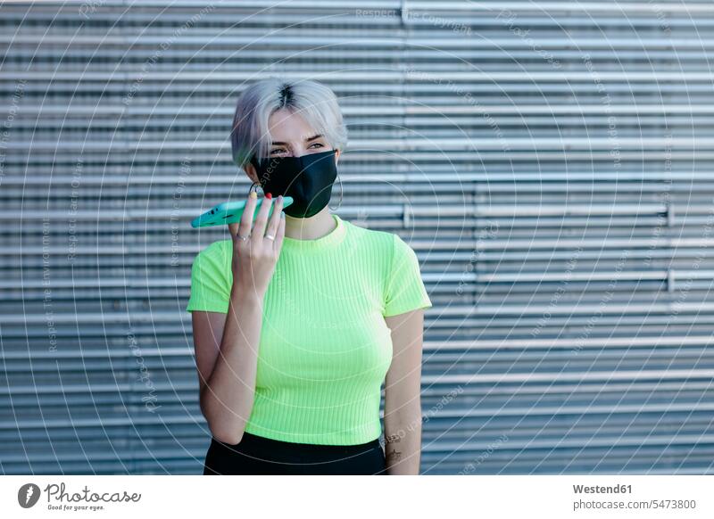 Blond woman standing in front of metal background, wearing face mask, using smartphone metallic metals telecommunication phones telephone telephones cell phone