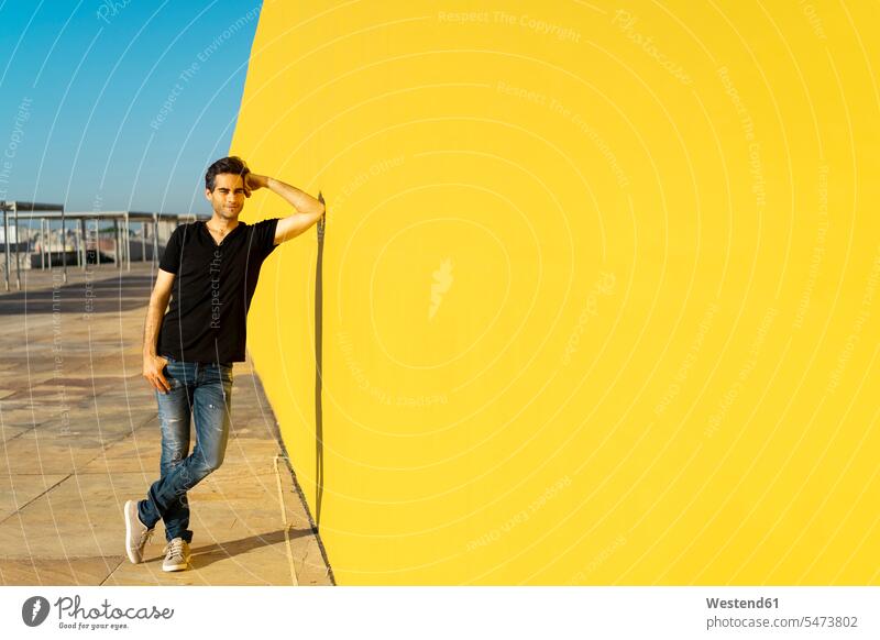 Man leaning against yellow wall, smiling, thinking standing man men males walls Adults grown-ups grownups adult people persons human being humans human beings