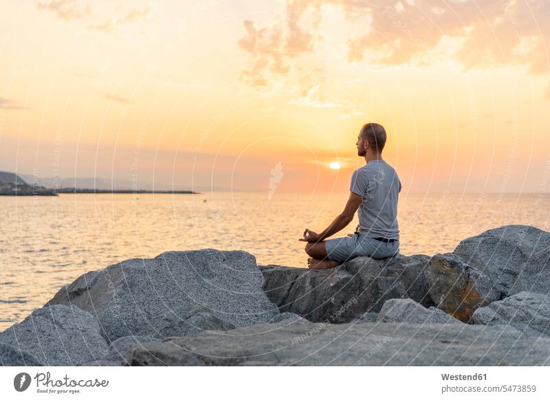 Spain. Man meditating during sunrise on rocky beach rocks meditate yoga meditation meditations man men males sitting Seated ocean mindfulness aware awareness