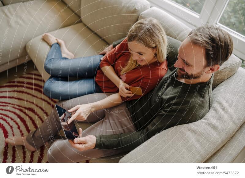 Couple sitting at home on couch, using digital tablet human human being human beings humans person persons caucasian appearance caucasian ethnicity european 2