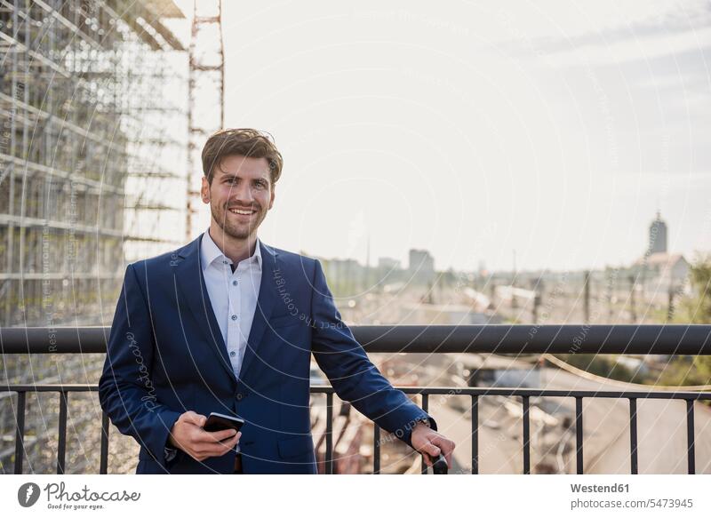 Portrait of smiling businessman standing on bridge in the city holding cell phone smile bridges Businessman Business man Businessmen Business men town cities