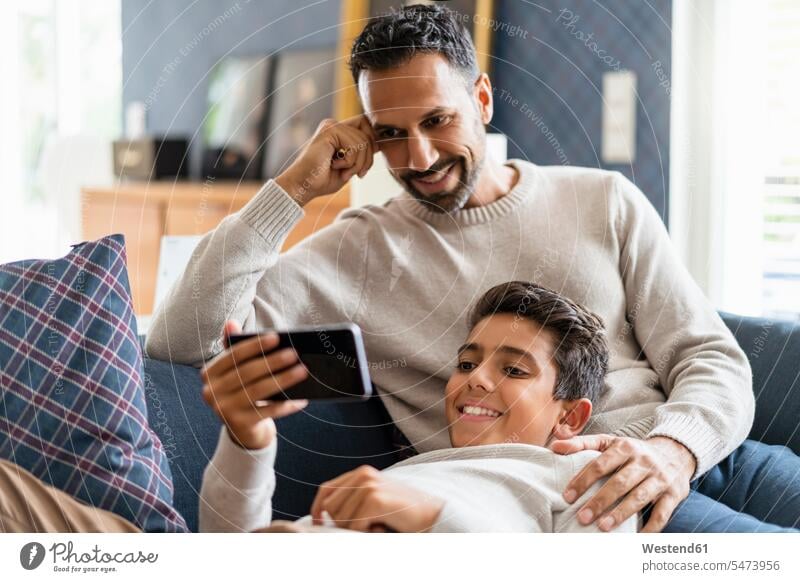 Smiling father and son using smartphone on couch in living room jumper sweater Sweaters couches settee settees sofa sofas telecommunication phones telephone