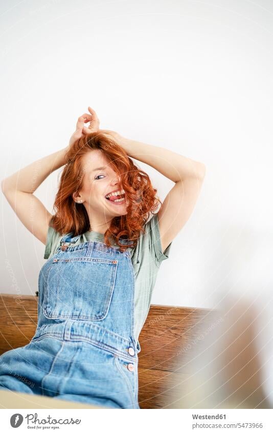 Cheerful redhead woman sitting with arms raised against white wall at home raising arms arm up arm raised arms up Hands Raised Hand Up Hands Up activity