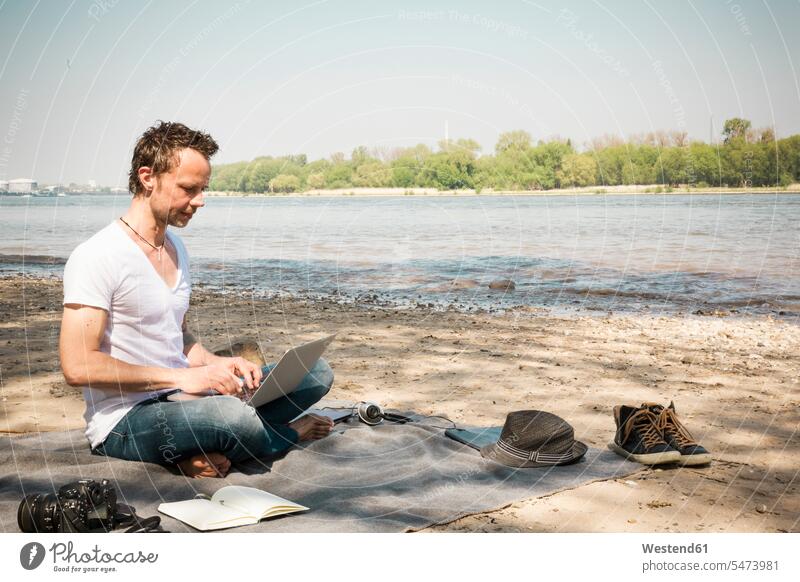 Man sitting on blanket at a river using laptop Laptop Computers laptops notebook Seated man men males Blanket Blankets beach beaches computer computers Adults