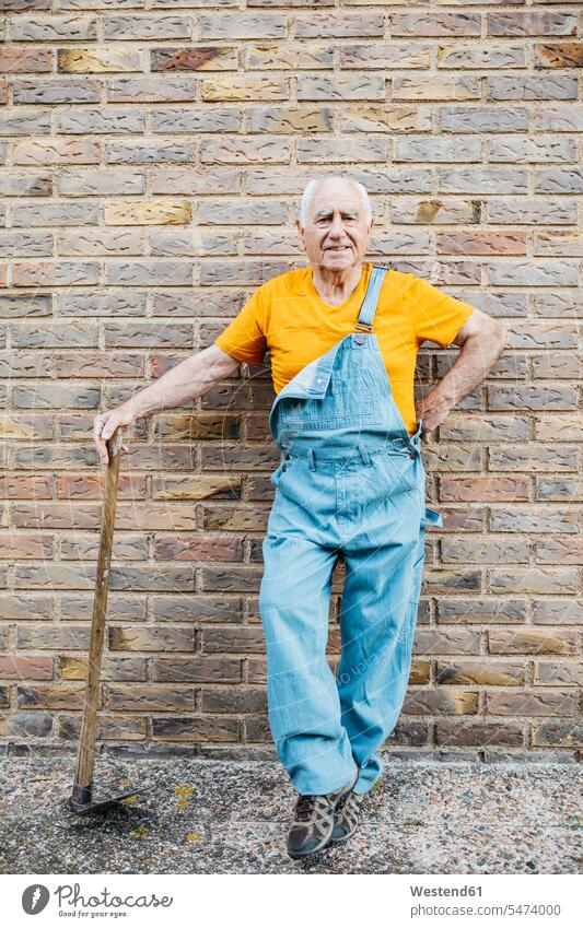 Senior man in denim overall with garden tool looking at camera leaning on a wall gardener gardeners looking to camera looking at the camera Eye Contact