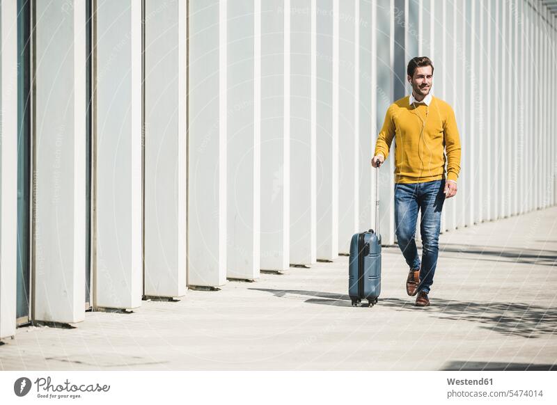 Young man with rolling suitcase and earphones in the city on the go touristic tourists business life business world business person businesspeople Business man
