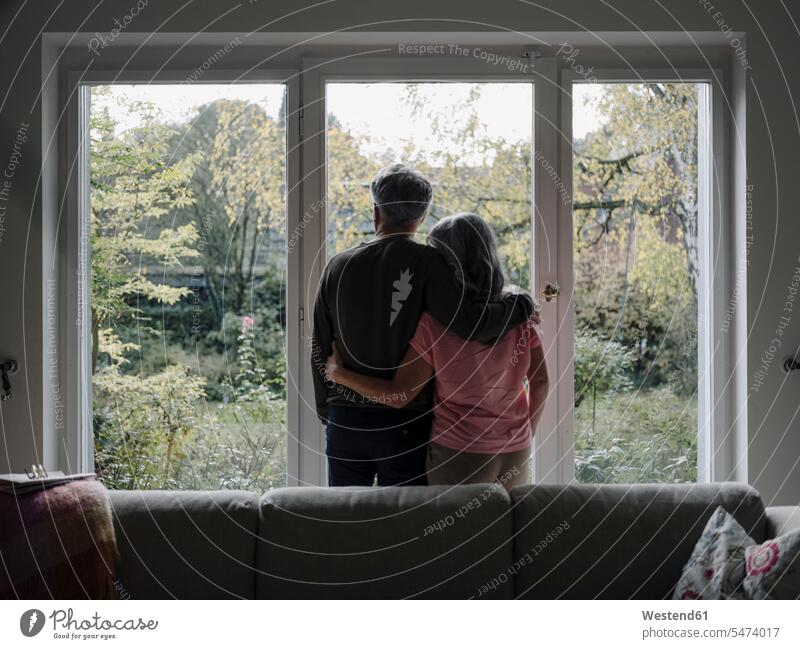 Rear view of senior couple looking out of window at home windows couches settee settees sofa sofas relax relaxing embrace Embracement hug hugging relaxation