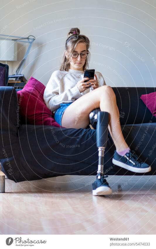 Young woman with leg prosthesis sitting on couch at home using smartphone human human being human beings humans person persons celibate celibates singles