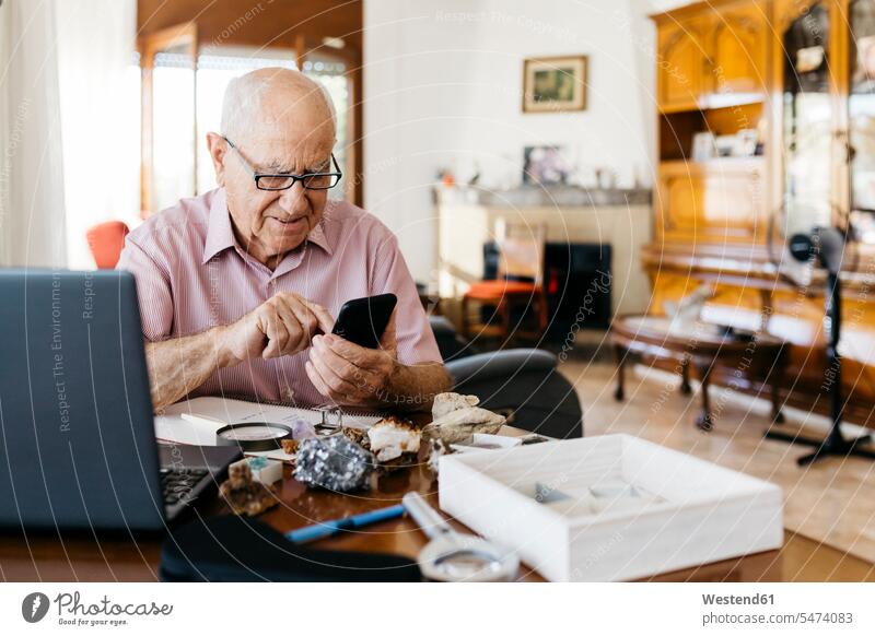 Elderly man using smart phone while doing research on mineral and fossil at home color image colour image indoors indoor shot indoor shots interior