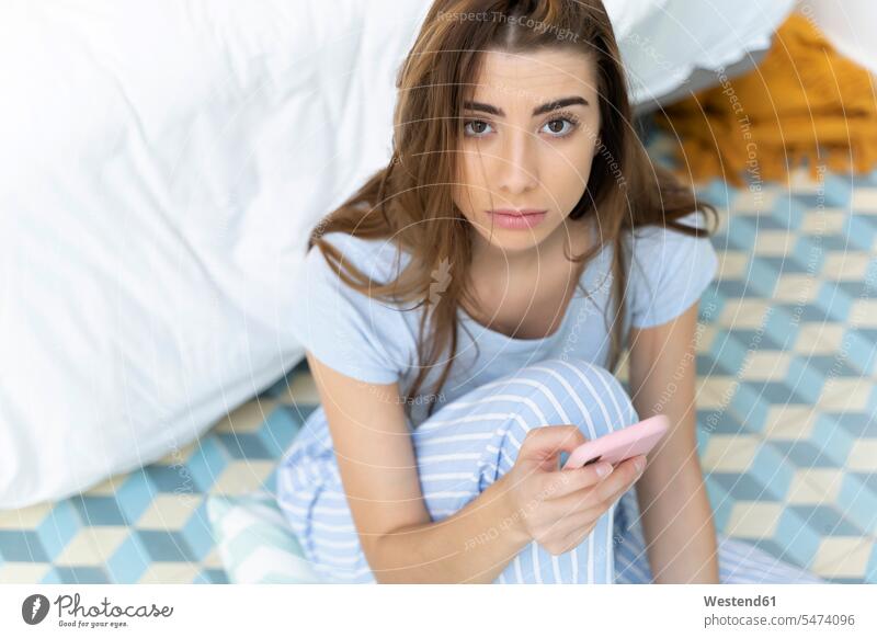 Portrait of young woman holding smartphone in bedroom Helplessness Bed - Furniture beds telecommunication phones telephone telephones cell phone cell phones