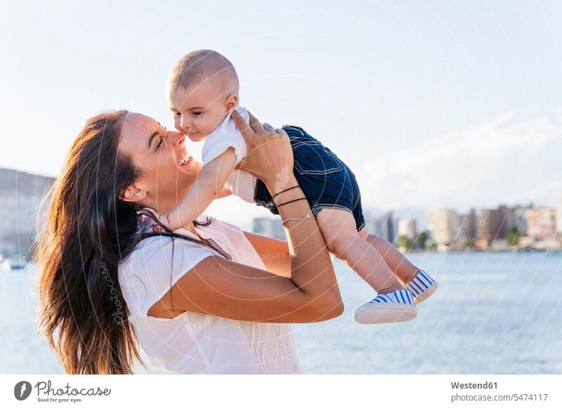 Smiling mother picking up baby son while standing against sea at sunset color image colour image Spain leisure activity leisure activities free time
