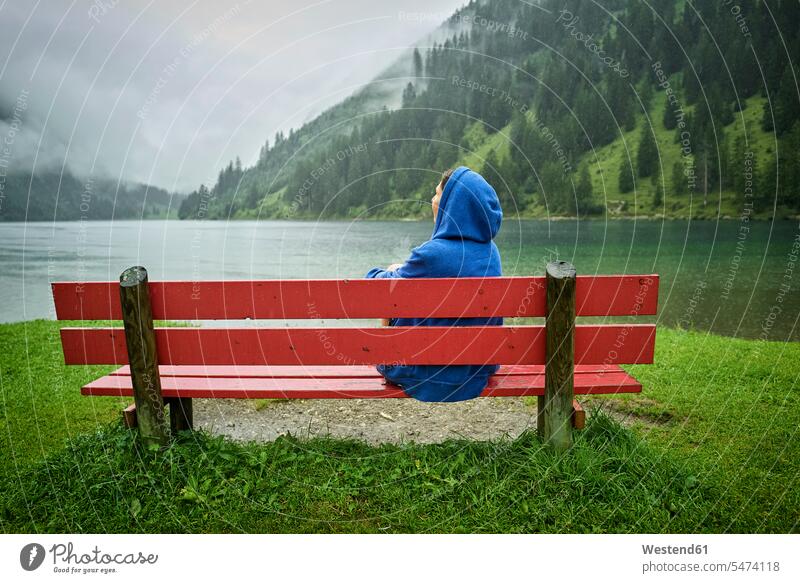 Woman wearing blue hood sitting on bench at lakeshore while looking at mountains color image colour image Austria outdoors location shots outdoor shot