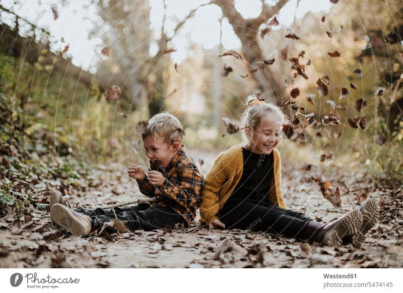 Cheerful girl and boy playing with dry fallen leaf while sitting on footpath in forest color image colour image outdoors location shots outdoor shot