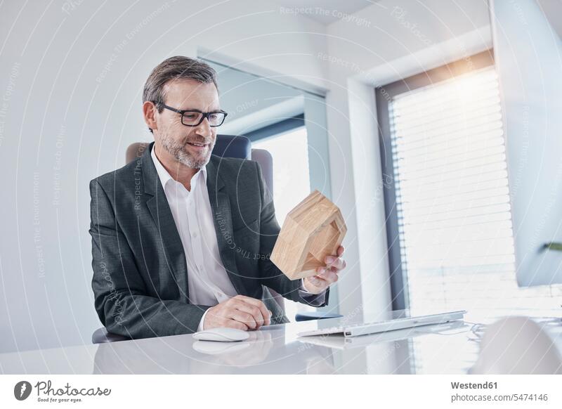 Smiling businessman at desk in office looking at architectural model desks offices office room office rooms Architectural Model smiling smile eyeing Businessman