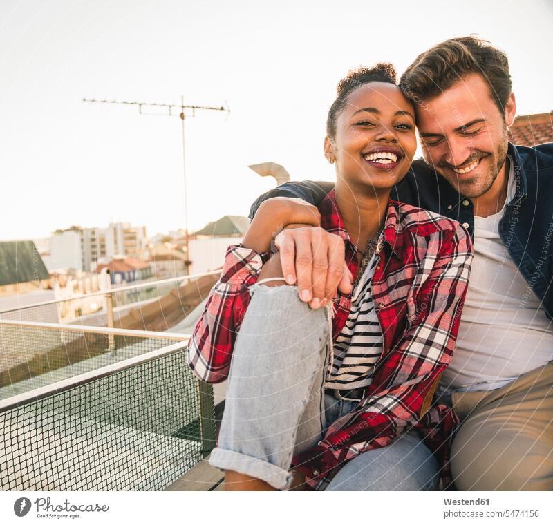 Happy affectionate young couple sitting on rooftop in the evening relax relaxing celebrate partying cuddle snuggle snuggling smile Seated embrace Embracement