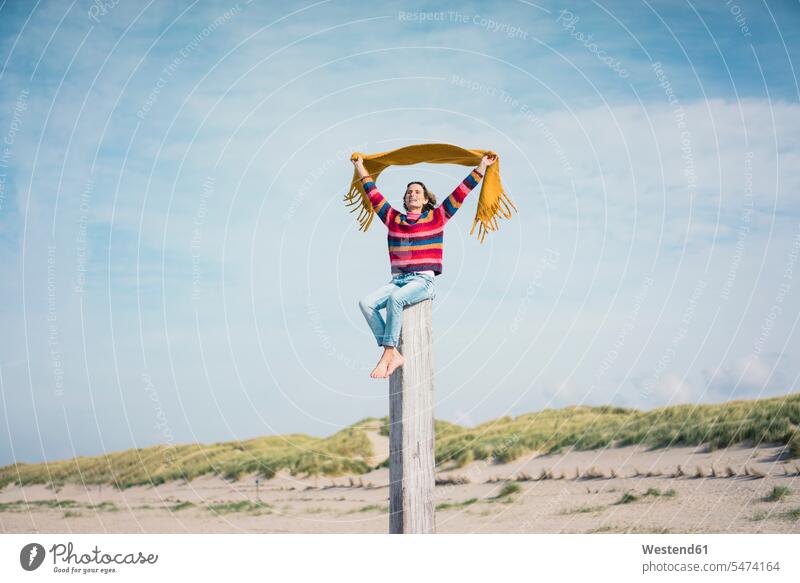Mature woman sitting on a wood pole on the beach, holding scarf in the wind scarfs scarves beaches Breeze windy wood post Wooden Poles flapping Seated