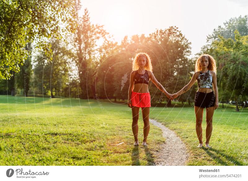 Twin sisters standing side by side in a park at backlight holding hands parks twin sister twin sisters paralell Juxtaposed in paralell Backlit back light