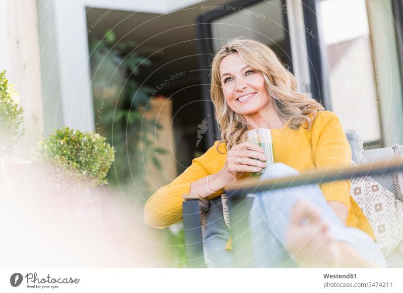 Portrait of smiling mature woman sitting on terrace with green smoothie Crockery Tableware Drinking Glass Drinking Glasses jumper sweater Sweaters chairs