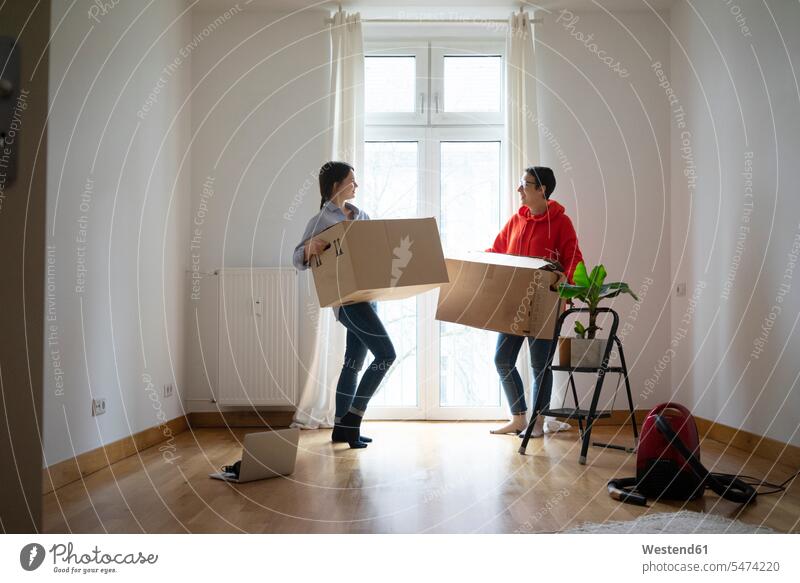 Young women moving into their new home, carrying cardboard boxes together moving house move Moving Home female friends packing case packing cases young women
