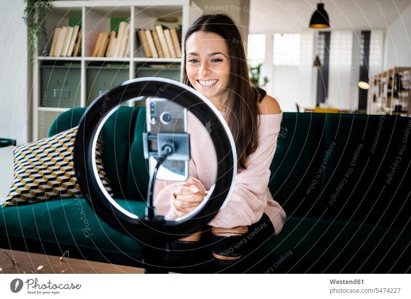Smiling beautiful female influencer sitting on sofa while vlogging through smart phone at home color image colour image indoors indoor shot indoor shots