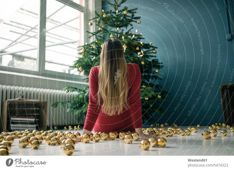 Back view of woman sitting on the floor with many golden Christmas baubles looking at Christmas tree Seated Floor Floors christmas bauble Christmas tree ball