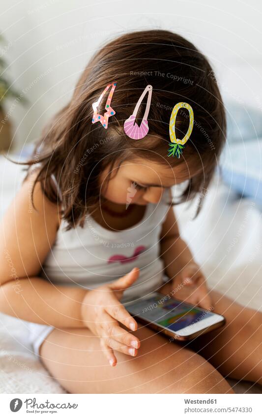 Little girl with three hair clips sitting on bed looking at smartphone Bed - Furniture beds cell phone cell phones Cellphone mobile mobile phones mobiles Seated