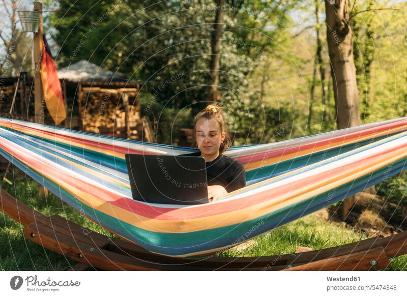Young woman studying on laptop while sitting in hammock against trees color image colour image outdoors location shots outdoor shot outdoor shots day