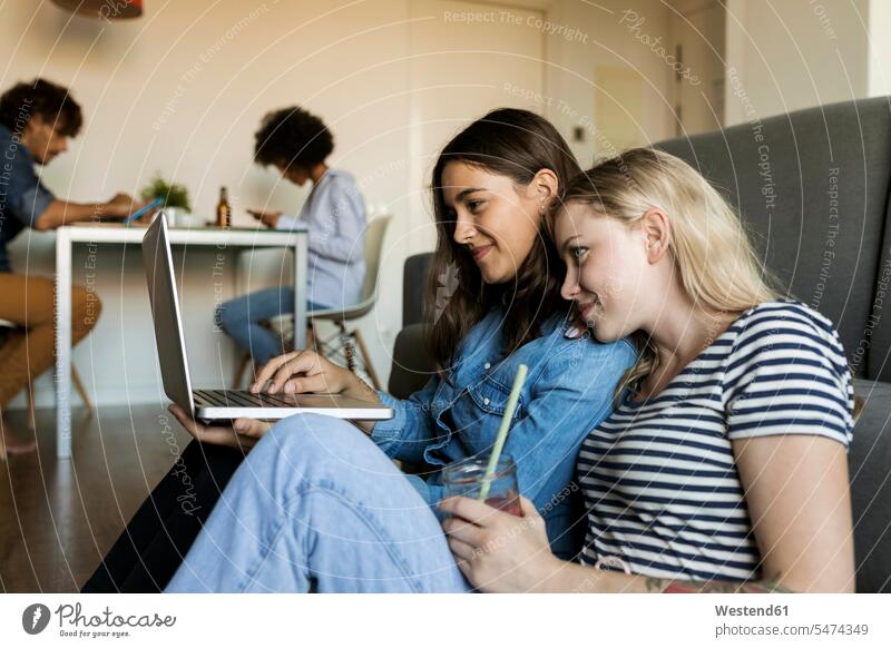 Two smiling young women sitting on floor sharing laptop with friends in background female friends share floors woman females Seated smile Laptop Computers