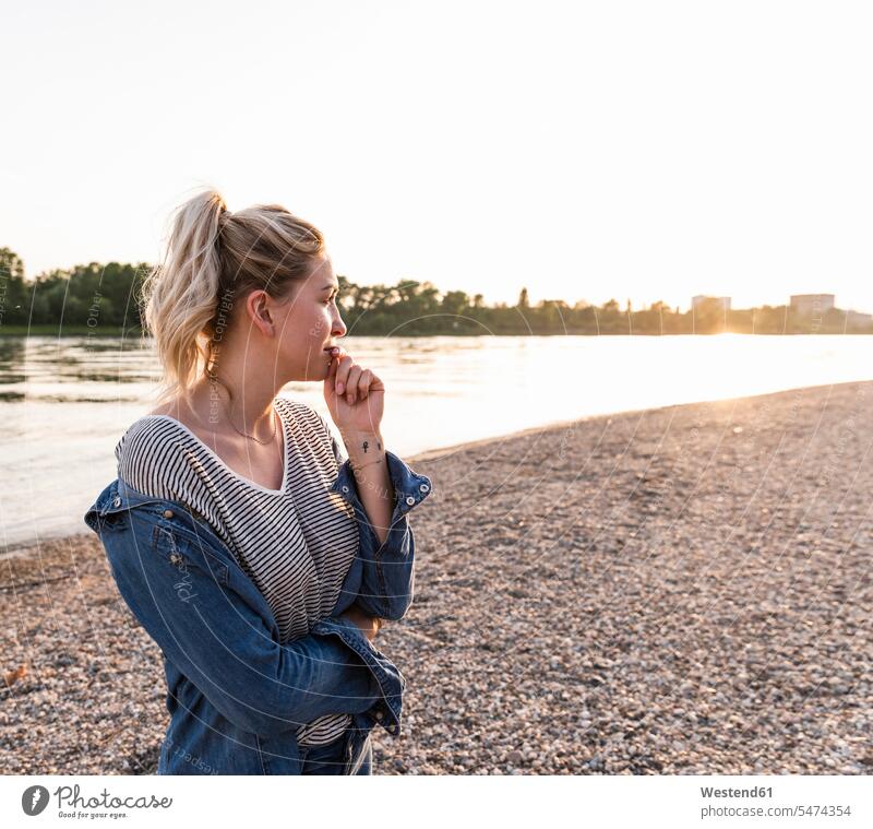 Young blond woman waiting on riverside in the evening pensive thoughtful Reflective contemplative standing riverbank females women looking sideways