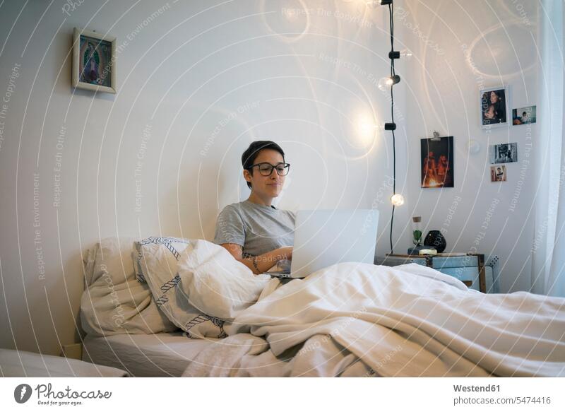 Young woman sitting in bed, using laptop looking eyeing watching looking at using a laptop Using Laptops young women young woman home at home Seated reading